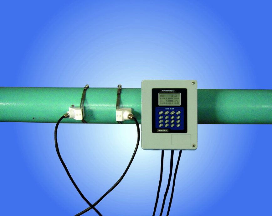A pair of ultrasonic transducers strapped to a green pipe, cabled to a wall mounted flow transmitter with a keypad and display