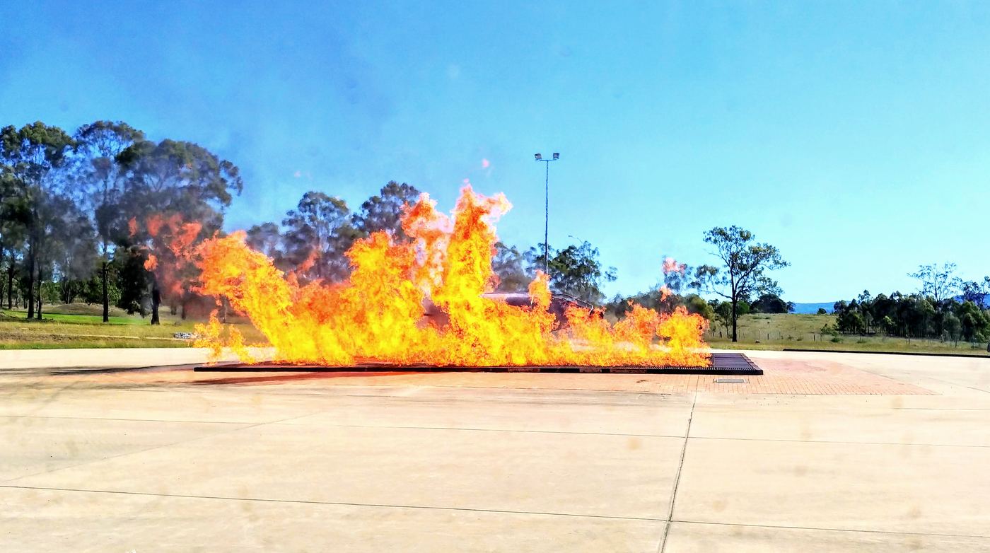 A live fire burning across a concrete training pad under a clear sky, with bushland behind