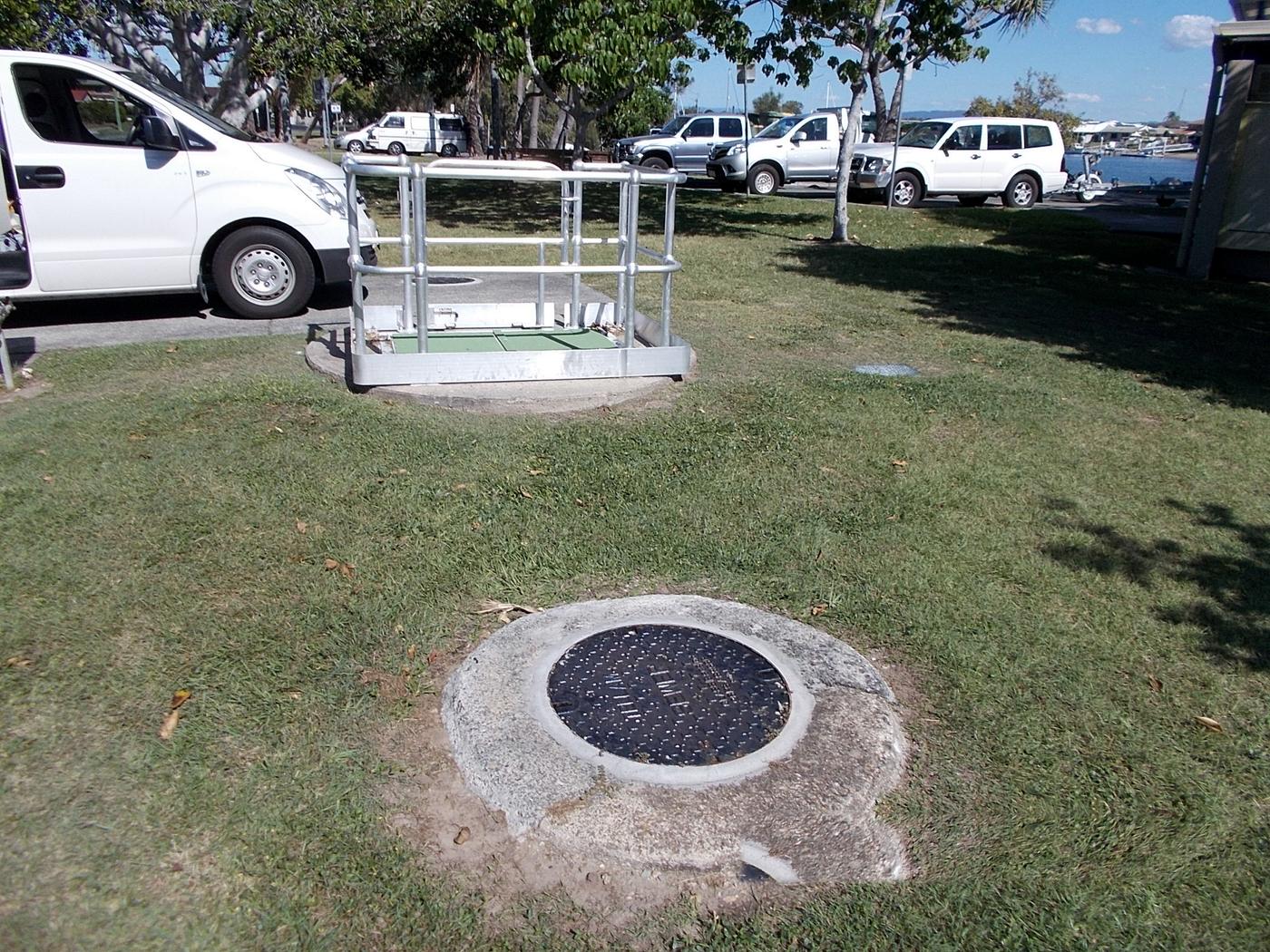 A sewer maintenance hole in a park with a safety barrier set up beside it and the EDS van parked nearby